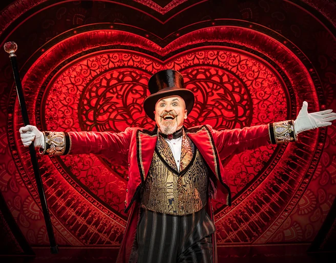 Moulin Rouge! The musical Ein strahlender Schauspieler im eleganten, roten Frack, mit Zylinder und Stock, vor leuchtendem Herzmotiv.A radiant actor in an elegant red tailcoat, with top hat and cane, in front of a glowing heart motif.