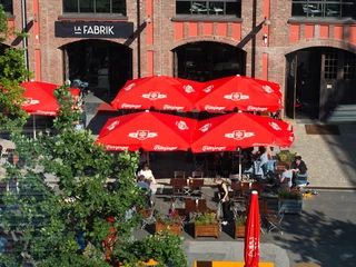 La Fabrik (2) Außengastronomie mit Schirmen und mehreren Sitzgruppen. Dahinter befindet sich ein Gebäude aus Ziegelsteinen, auf dem "La Fabrik" steht.