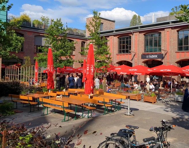 La Fabrik (1) Außenterrasse mit roten quadratischen Schirmen und Sitzgruppen, die von einigen Menschen besucht sind. Dahinter befindet sich ein großes Gebäude aus Ziegelsteinen.