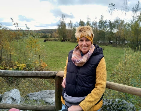 Outdoor-Relax Training mit Anja Lindemann Anja Lindemann lächelt in herbstlicher Kleidung an einem Holzzaun vor grüner Wiese mit Bäumen.