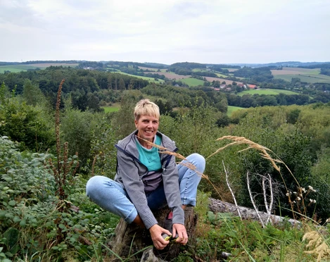 Outdoor-Relax Training mit Anja Lindemann Eine Person sitzt im Freien auf einem Baumstamm, umgeben von grünen Feldern und hügeliger Landschaft.