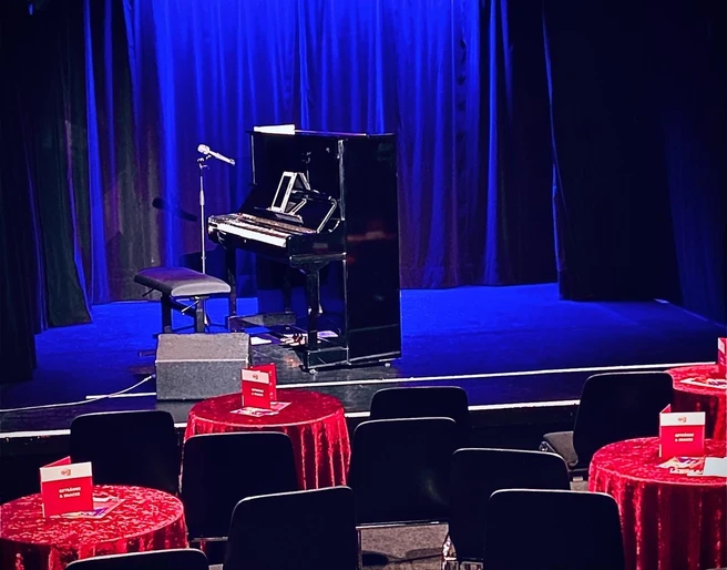Theater509 / Bürgerhaus Stollwerck Ein gemütlicher Theatersaal mit roten Tischdecken, Stühlen, einem Piano und einem stehenden Mikrofon.A cozy theater hall with red tablecloths, chairs, a piano and a standing microphone.