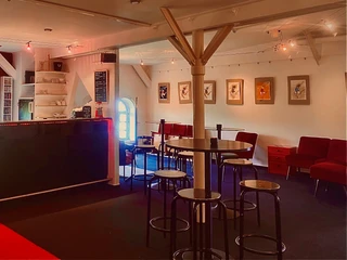 Theater509 / Bürgerhaus Stollwerck Gemütlicher Innenraum eines Cafés mit Holztischgruppe, roter Couch und Bildergalerie an der Wand.Cozy interior of a café with a group of wooden tables, a red couch and a picture gallery on the wall.