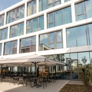 Außenansicht des Hotels mit großer Glasfront. Im Eingangsbereich Stühle und Tische mit Sonnenschirmen