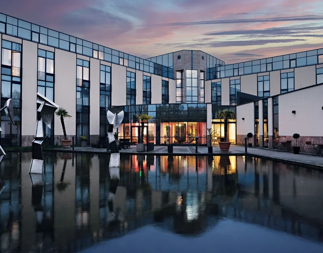 Außenansicht des Four Points Flex by Sheraton Ratingen Düsseldorf Airport Moderne Hotelfassade mit Glasflächen, Skulpturen und Wasserbecken bei Abenddämmerung in Ratingen.