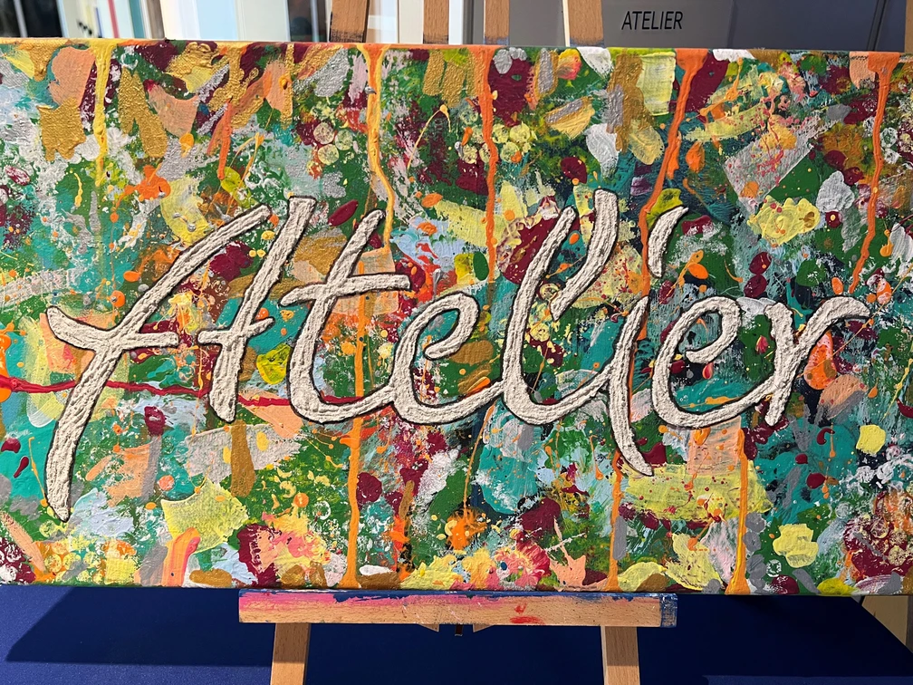 Atelier Vielseitig gestaltete Leinwand mit dem Schriftzug "Atelier" vor einer Staffelei, farbenreich bemalt.