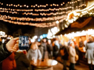 Weihnachtsdorf im Lichterglanz Hand, die eine Tasse Glühwein hält im Fokus und das beleuchtete Einbecker Weihnachtsdorf dahinter.