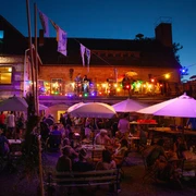 KulturOrt Heymannbaude Ein lebhaftes Abendfest im Freien mit bunten Lichtern, Menschen unter Sonnenschirmen und einer Terrasse mit Musikern vor einem beleuchteten Gebäude.