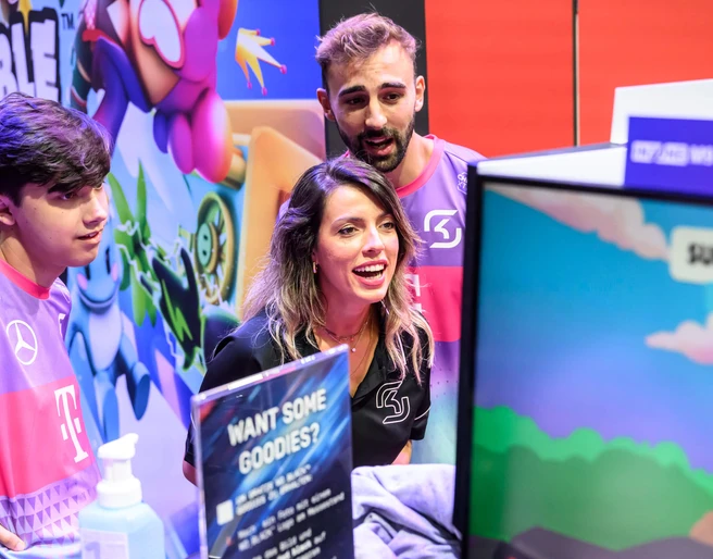 gamescom Eine dynamische Szene bei der Gamescom: Drei Personen in lila T-Shirts interagieren begeistert mit einem Videospiel auf einem Bildschirm.A dynamic scene at Gamescom: three people in purple T-shirts interact enthusiastically with a video game on a screen.
