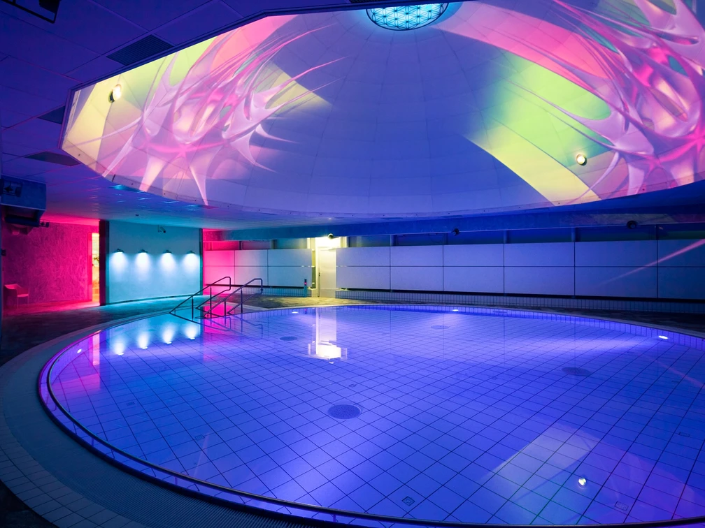 Liquid Sound Tempel Toskana Therme Beleuchtetes Hallenbad mit rundem Becken, blauen und pinken Lichtern sowie bunten Projektionen an der Decke, moderne und entspannte Atmosphäre.