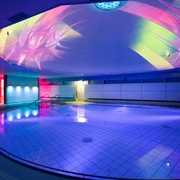 Liquid Sound Tempel Toskana Therme Beleuchtetes Hallenbad mit rundem Becken, blauen und pinken Lichtern sowie bunten Projektionen an der Decke, moderne und entspannte Atmosphäre.