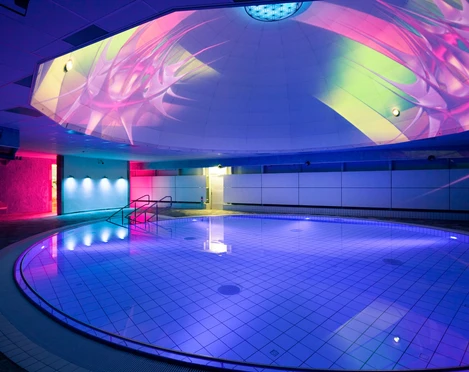 Liquid Sound Tempel Toskana Therme Beleuchtetes Hallenbad mit rundem Becken, blauen und pinken Lichtern sowie bunten Projektionen an der Decke, moderne und entspannte Atmosphäre.