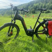 Fahrradmodell mit 625 Wh-Akku.jpg Schwarzes E-Bike der Marke Haibike mit roter Fahrradtasche steht auf einer grünen Wiese vor bewaldeter Hügellandschaft bei sonnigem Wetter.
