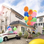 Kristallhotel FETTEHENNE in Leverkusen Ein modernes Hotelgebäude mit bunten Luftballons und einem Oldtimer, umgeben von klarer, blauer Himmel.