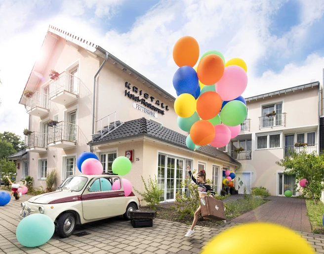 Kristallhotel FETTEHENNE in Leverkusen Ein modernes Hotelgebäude mit bunten Luftballons und einem Oldtimer, umgeben von klarer, blauer Himmel.