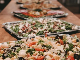 Verschiedene reichhaltig belegte Salatplatten stehen auf einem langen Holztisch, Hintergrund unscharf.
