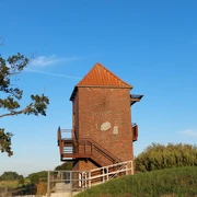 Landschaftsfenster Tange.jpg Das Bild zeigt einen Backsteinturm mit einem spitzen, roten Ziegeldach. Der Turm ist mit hölzernen Treppen und Geländern umgeben, die Besucher zur Aussichtsplattform führen. Links im Vordergrund steht ein großer Baum, und im Hintergrund erstreckt sich die Landschaft unter einem klaren blauen Himmel. Der Turm befindet sich in einer grünen Umgebung, die auf die ländliche Idylle der Region hindeutet.