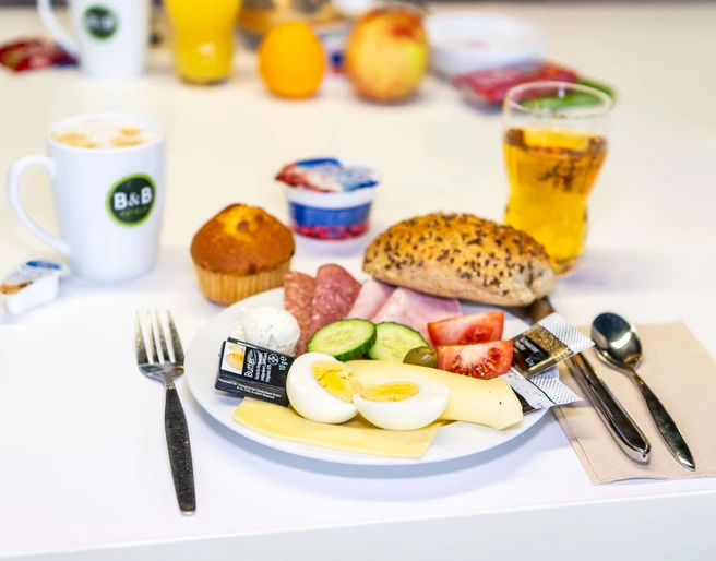 Frühstück im B&B HOTEL Düsseldorf-Hilden Ein Teller mit frischem Frühstück: Aufschnitt, belegtes Brötchen, Heißgetränk und Saft im Hintergrund.