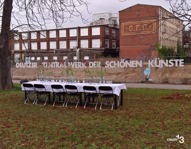 raum13  Eine lange, festlich gedeckte Tafel steht unter freiem Himmel vor einer alten Backsteinfabrik.A long, festively laid table stands in the open air in front of an old brick factory.