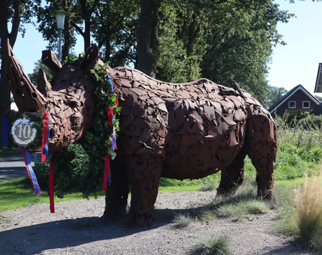 Nashorn-Spohle-DerWiefelsteder.JPG Das Bild zeigt eine lebensgroße Statue eines Nashorns in einer ländlichen Umgebung. Die Statue besteht aus rostigem Metall und trägt einen Kranz um den Hals, was auf eine Feier oder ein lokales Event hindeutet. Im Hintergrund sind Bäume und ein Wohnhaus zu sehen.