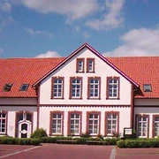 RathausKirchstraße10.jpg Das Bild zeigt ein zweistöckiges Gebäude mit leuchtend roten Dachziegeln und weißen Wänden. Die Fassade hat mehrere symmetrisch angeordnete Fenster mit roten Backsteinrahmen.