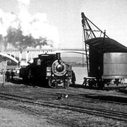 Historische Dampflok vor Eisenbahnbrücke am Fluss in Schwarz-Weiß, Dampf steigt aus dem Schornstein empor.