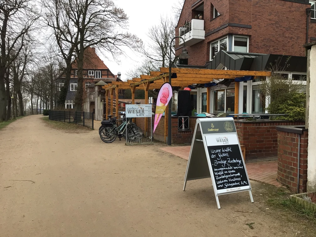 Garbes Weser-Café Ein Weg führt an der Terrasse von Garbes Weser-Café entlang, umgeben von Bäumen und einem Gebäude.