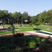 minigolfplatz.jpg Grüner Minigolfplatz mit mehreren Bahnen, umgeben von Bäumen; Menschen spielen bei sonnigem Wetter.
