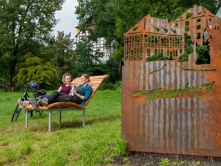 rastplatz_tonenburg_Henning_Orendt (8).jpg Zwei Frauen auf einer Landschaftsliege mit einer Cortenstahlarbeit der Tonenburg und der Tonnenburg im Hintergrund