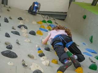 Sicherungsautomat im Kletterbereich Ein Kind klettert eine bunte Kletterwand hoch, gesichert mit einem blauen Klettergurt; die Atmosphäre wirkt konzentriert und sportlich.