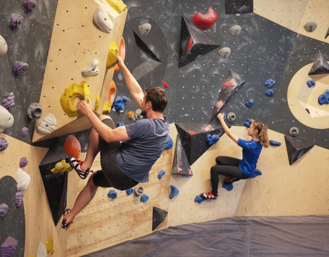 Boulderbereich in der YOYO-Kletterhalle Zwei Personen klettern an einer Indoor-Kletterwand mit bunten Griffen; der Mann trägt ein graues Shirt, die Frau ein blaues.