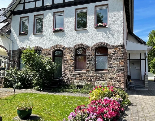 fewo_hermanns_aussenansicht.jpg Zweistöckiges Fachwerkhaus mit Steinbasis, blühenden Beeten und gepflegter Rasenfläche im Vordergrund.