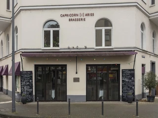 Brasserie Capricorn i aries  Ansicht auf die helle Fassade des Restaurants mit zwei Kreidetafeln, die das Menü präsentieren. View of the bright facade of the restaurant with two chalkboards presenting the menu.