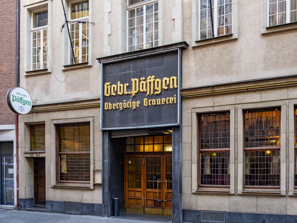 Brewery Päffgen Eingang zum Brauhaus durch eine rustikale Holztür, darüber ein mit goldenen Buchstaben verziertes Schild.The entrance to the brewery is through a rustic wooden door, with a sign above it decorated with golden letters.