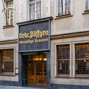 Brewery Päffgen Eingang zum Brauhaus durch eine rustikale Holztür, darüber ein mit goldenen Buchstaben verziertes Schild.The entrance to the brewery is through a rustic wooden door, with a sign above it decorated with golden letters.