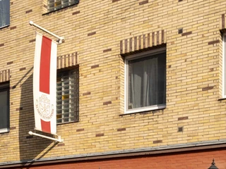 Brewery Schreckenskammer Ansicht der Fassade des Brauhauses Schreckenskammer in Köln. Das Gebäude zeigt eine einstöckige rote Backsteinfront mit dem Schriftzug "Biergarten" in einem Fenster und drei Wandlaternen, die an der Fassade befestigt sind. Eine rot-weiße Fahne mit dem Wappen des Brauhauses hängt an der oberen Etage des gelb-rot gemusterten Hauses.View of the facade of the Brauhaus Schreckenskammer in Cologne. The building features a single-story red brick front with the inscription "Biergarten" displayed in one of the windows and three wall-mounted lanterns on the facade. A red and white flag bearing the brewery's emblem hangs from the upper floor of the building, which is decorated in a yellow and red patterned design.