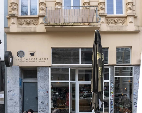 The Coffee Gang Die Fassade eines eleganten Altbaus mit ornamentierten Fenstern beherbergt das Café „The Coffee Gang“. Im Vordergrund stehen Holztische und Stühle, einige Gäste unterhalten sich im Freien. The facade of an elegant historic building with ornately decorated windows houses the café "The Coffee Gang". Wooden tables and chairs stand in the foreground, with some guests chatting outside.