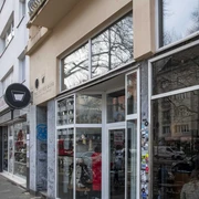 The Coffee Gang Eingang des modernen Cafés "The Coffee Gang" in Köln. Eine breite Glasfront, flankiert von Graffititags auf Naturstein, lässt viel Licht in den Innenraum fallen. Außen stehen Fahrräder geparkt, während das Umfeld von Wohngebäuden mit schlichter Architektur geprägt ist.Entrance to the modern café "The Coffee Gang" in Cologne. A wide glass facade, flanked by graffiti tags on natural stone, allows ample light into the interior space. Bicycles are parked outside, while the surroundings are characterized by residential buildings with simple architecture.