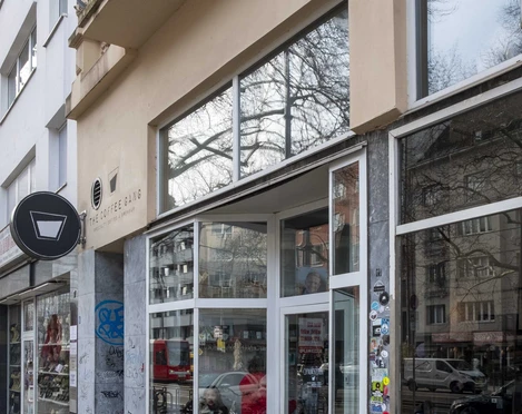 The Coffee Gang Eingang des modernen Cafés "The Coffee Gang" in Köln. Eine breite Glasfront, flankiert von Graffititags auf Naturstein, lässt viel Licht in den Innenraum fallen. Außen stehen Fahrräder geparkt, während das Umfeld von Wohngebäuden mit schlichter Architektur geprägt ist.Entrance to the modern café "The Coffee Gang" in Cologne. A wide glass facade, flanked by graffiti tags on natural stone, allows ample light into the interior space. Bicycles are parked outside, while the surroundings are characterized by residential buildings with simple architecture.
