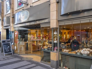 Cafe Printen Schmitz Die Außenseite eines Cafés mit dem Namen "Printen Schmitz" in Köln, das eine Vielzahl von Gebäcken und Süßwaren in den Schaufenstern präsentiert. Vor dem Eingang sind Tische und Stühle aufgestellt. Menschen sind zu sehen, die im Inneren des Cafés beschäftigt sind.The exterior of a café called ‘Printen Schmitz’ in Cologne, displaying a variety of pastries and confectionery in the shop windows. Tables and chairs are set up in front of the entrance. People can be seen busy inside the café.