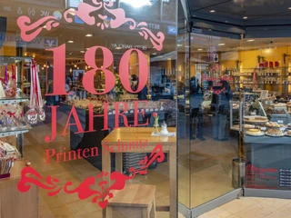 Cafe Printen Schmitz Im Schaufenster des Cafés Printen Schmitz reflektiert die Aufschrift "180 Jahre" die traditionsreiche Jubiläumsfeier, während drinnen vielfältige Backwaren einladend präsentiert werden.In the window of Café Printen Schmitz, the inscription ‘180 years’ reflects the traditional anniversary celebrations, while inside, a wide variety of baked goods are invitingly displayed.