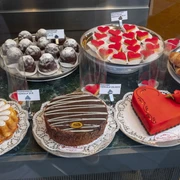 Cafe Printen Schmitz Eine einladende Vitrine eines Cafés mit einer Auswahl an kunstvoll dekorierten Süßwaren. Zu sehen sind eine Vielzahl von Kuchen und Torten, darunter eine auffällige herzförmige Torte mit einer roten Verzierung, die auf einer weißen Tortenspitze präsentiert wird.An inviting café display case with a selection of artfully decorated confections. On display are a variety of cakes and pies, including a striking heart-shaped pie with red decoration, presented on a white doily.