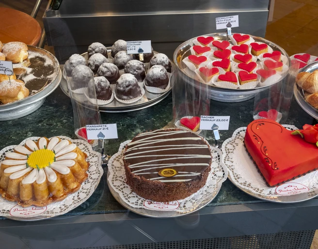 Cafe Printen Schmitz Eine einladende Vitrine eines Cafés mit einer Auswahl an kunstvoll dekorierten Süßwaren. Zu sehen sind eine Vielzahl von Kuchen und Torten, darunter eine auffällige herzförmige Torte mit einer roten Verzierung, die auf einer weißen Tortenspitze präsentiert wird.An inviting café display case with a selection of artfully decorated confections. On display are a variety of cakes and pies, including a striking heart-shaped pie with red decoration, presented on a white doily.