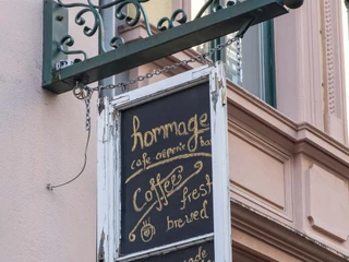 Cafe Hommage  Eine Kreidetafel im Shabby Chic Stil ist an einem Metallgestell an der Hausfassade befestigt.A chalkboard in a shabby chic style is attached to a metal frame on the house facade.