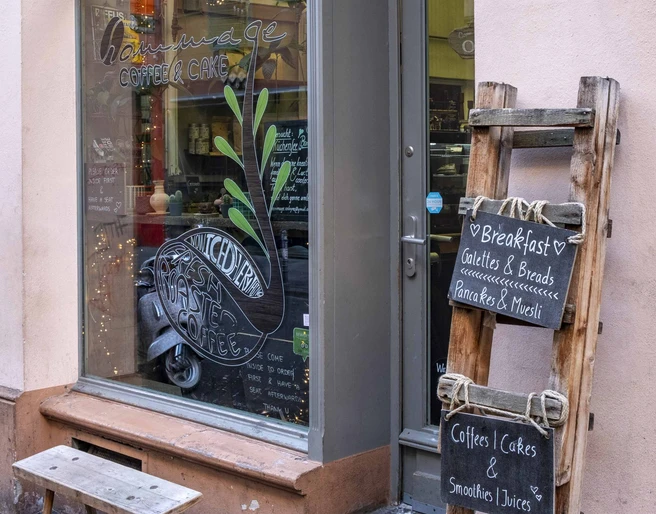 Cafe Hommage  Einladendes Fenster mit kleiner Holzbank davor, daneben eine Kreidetafel an einer Holzleiter befestigt.Inviting window with a small wooden bench in front of it, next to it a chalk board attached to a wooden ladder.