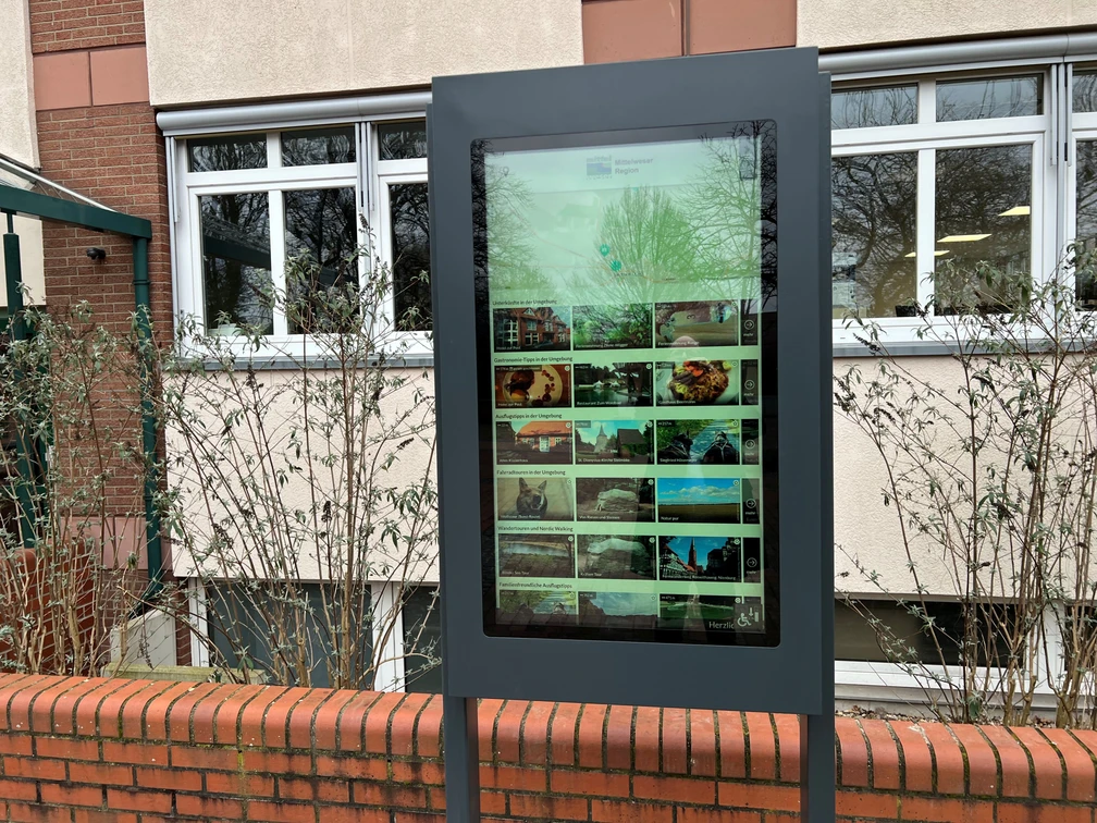 Infoterminal Steimbke  Digitales Infoterminal an einer Ziegelmauer vor einem Gebäude, von Sträuchern umgeben.