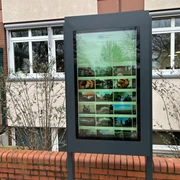 Infoterminal Steimbke Digitales Infoterminal an einer Ziegelmauer vor einem Gebäude, von Sträuchern umgeben.