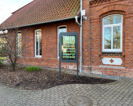 Infoterminal Stolzenau  Infotafel in Stolzenau vor rotem Backsteingebäude, flankiert von gepflastertem Weg und Beeten.