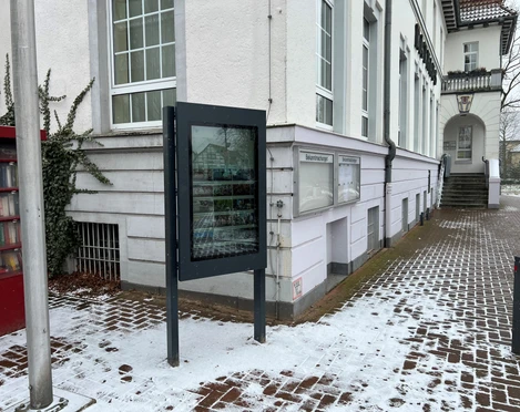 Infoterminal Uchte  Ein Infoterminal steht vor einem schneebedeckten Gehweg neben einem historischen Gebäude mit weißen Wänden.