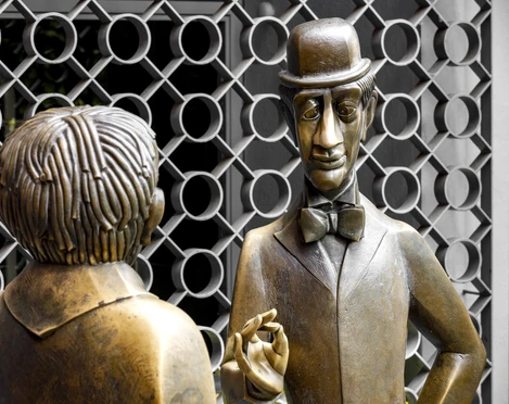 Tünnes-und-Schäl-Denkmal Das Bild zeigt die berühmten Bronze-Figuren "Tünnes und Schäl" am Willy-Millowitsch-Platz in Köln. Beide Figuren stehen einander gegenüber; Tünnes trägt eine Mütze und Schäl einen Zylinder. Im Hintergrund ist ein kunstvoll geschmiedetes Gitter zu sehen.The picture shows the famous bronze figures "Tünnes and Schäl" at Willy-Millowitsch-Platz in Cologne. Both figures are standing opposite each other; Tünnes is wearing a cap and Schäl a top hat. An artistically forged grille can be seen in the background.